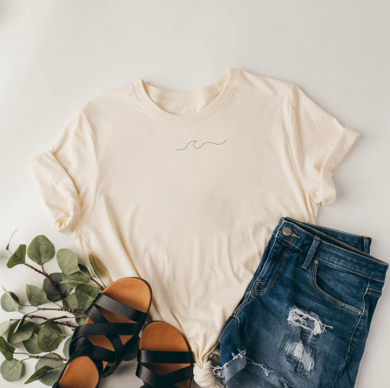 flat lay of cream shirt with calm wave logo centered under neckline, shoes and jeans laying flat on top to compliment shirt.